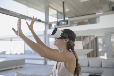 Smiling woman using virtual reality simulator glasses with arms raised in modern, luxury home showcase living room - HOXF01290