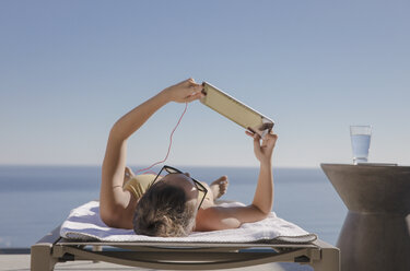 Frau, die sich sonnt und ein digitales Tablet auf einem Liegestuhl auf einer sonnigen Terrasse mit Meerblick benutzt - HOXF01292