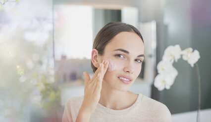 Portrait brunette woman applying face moisturizer to cheek - HOXF01303