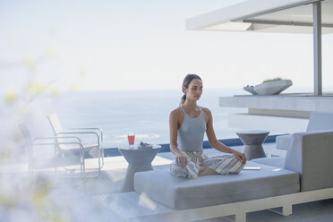 Serene woman meditating in lotus position on modern, luxury home showcase exterior patio sofa with ocean view - HOXF01322