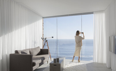 Frau mit Fotohandy fotografiert sonnigen Blick auf das Meer von modernen, luxuriösen Haus Schaufenster Innenraum Wohnzimmer - HOXF01328
