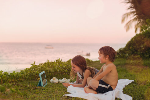 Junge und Mädchen Bruder und Schwester beobachten Video auf digitalen Tablet im Gras mit Blick auf das Meer - HOXF01372