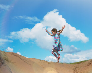 Exuberant girl jumping for joy on beach hill below sunny blue sky with clouds - HOXF01384