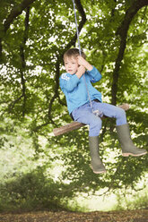 Boy in wellies swinging on tree rope swing - HOXF01417