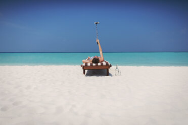 Woman laying, sunbathing and taking selfie with selfie stick on lounge chair on sunny tropical beach - HOXF01427