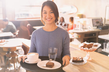 Porträt einer lächelnden Kellnerin, die ein Tablett mit Cappuccino, Brownie und Wasser in einem Cafe trägt - HOXF01492