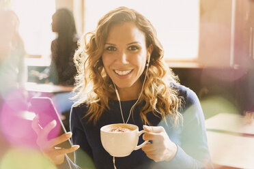 Porträt einer lächelnden Frau mit Kopfhörern, die Musik auf einem mp3-Player hört und Cappuccino in einem Café trinkt - HOXF01517