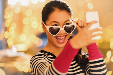 Playful Chinese woman wearing heart-shape glasses taking selfie with camera phone - HOXF01568