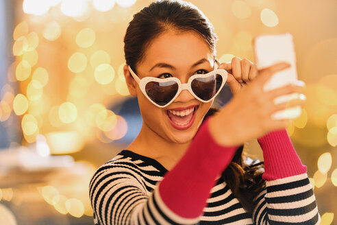 Verspielte chinesische Frau mit herzförmiger Brille, die ein Selfie mit einem Fotohandy macht - HOXF01568
