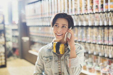 Lächelnde junge Frau mit Kopfhörern, die in einem Lebensmittelgeschäft mit ihrem Handy telefoniert - HOXF01614