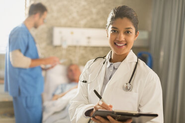 Portrait smiling female doctor making rounds, taking notes on medical record clipboard in hospital room - HOXF01716