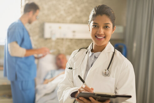 Porträt einer lächelnden Ärztin bei der Visite, die sich Notizen auf einem Klemmbrett in einem Krankenhauszimmer macht - HOXF01716