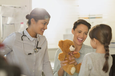 Smiling female pediatrician and mother showing teddy bear to girl patient in examination room - HOXF01733