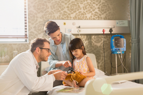 Männlicher Arzt, der ein Otoskop an einem Teddybär eines Mädchens im Krankenhauszimmer benutzt, lizenzfreies Stockfoto