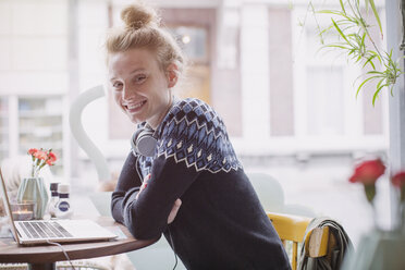 Porträt einer lächelnden jungen Frau mit Kopfhörern am Laptop im Caféfenster - HOXF01779