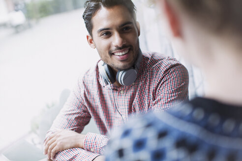 Lächelnder junger Mann mit Kopfhörern im Gespräch mit dem Fenster eines Cafés - HOXF01788
