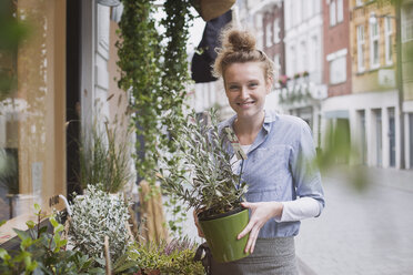 Porträt einer lächelnden Blumenhändlerin, die eine Topfpflanze im Schaufenster hält - HOXF01790