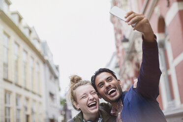 Enthusiastic, laughing young couple taking selfie in city - HOXF01800