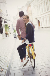 Porträt einer lächelnden Frau, die auf der Rückseite des Fahrrads ihres Freundes auf einer Stadtstraße fährt - HOXF01817