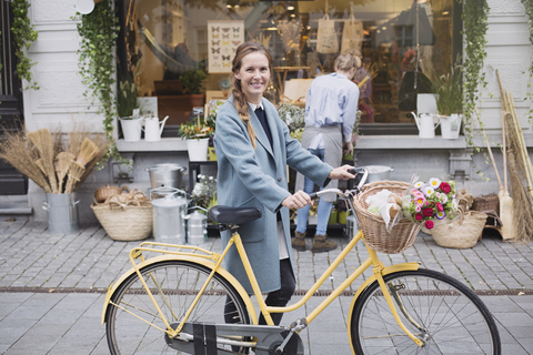 Porträt lächelnde Frau mit Fahrrad und Blumen im Korb vor einem Geschäft, lizenzfreies Stockfoto