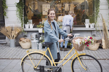 Porträt lächelnde Frau mit Fahrrad und Blumen im Korb vor einem Geschäft - HOXF01827