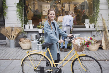 Portrait smiling woman walking bicycle with flowers in basket outside storefront - HOXF01827