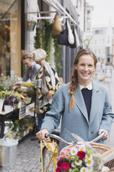 Portrait smiling woman walking bicycle with flowers in basket outside storefront - HOXF01831