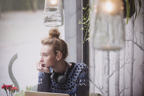 Nachdenkliche junge Frau mit Kopfhörern am Laptop, die aus dem Fenster eines Cafés schaut, lizenzfreies Stockfoto
