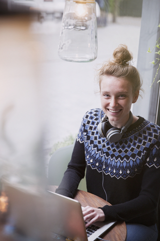Porträt einer lächelnden jungen Frau, die in einem Caféfenster einen Laptop benutzt, lizenzfreies Stockfoto