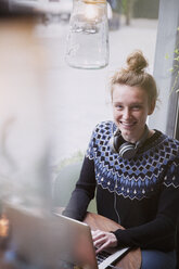 Porträt einer lächelnden jungen Frau, die in einem Caféfenster einen Laptop benutzt - HOXF01845
