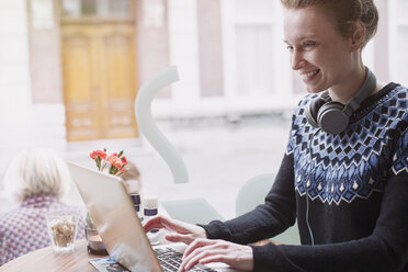 Smiling young woman with headphones using laptop in sidewalk cafe window - HOXF01856