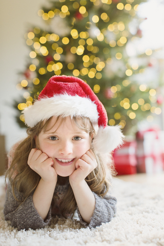 Porträt lächelndes Mädchen mit Weihnachtsmannmütze auf Teppich im Wohnzimmer mit Weihnachtsbaum, lizenzfreies Stockfoto