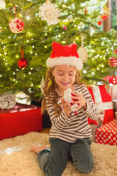 Smiling girl in Santa hat opening gift in front of Christmas tree - HOXF01874