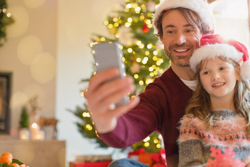 Lächelnder Vater und Tochter mit Weihnachtsmannmützen machen ein Selfie mit einem Fotohandy - HOXF01876