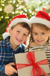 Porträt lächelnder Bruder und Schwester mit Weihnachtsmannmützen, die ein Weihnachtsgeschenk halten - HOXF01882
