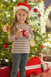 Porträt eines lächelnden Mädchens mit Weihnachtsmannmütze und Geschenk vor einem Weihnachtsbaum - HOXF01886