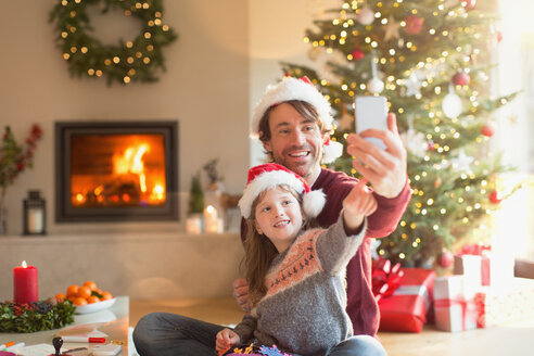 Vater und Tochter mit Weihnachtsmannmützen machen ein Selfie mit Fotohandy im Weihnachtswohnzimmer - HOXF01894