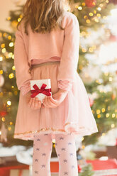 Mädchen in rosa Kleid hält Weihnachtsgeschenk hinter ihrem Rücken in der Nähe von Weihnachtsbaum - HOXF01901