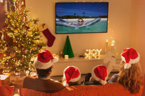 Familie mit Weihnachtsmannmützen sieht sich im Weihnachtswohnzimmer ein Sommerferienvideo im Fernsehen an - HOXF01947