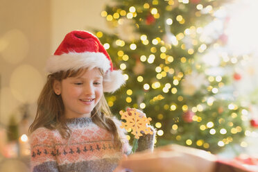 Mädchen mit Weihnachtsmannmütze hält Schneeflockenornament vor einem Weihnachtsbaum - HOXF01960