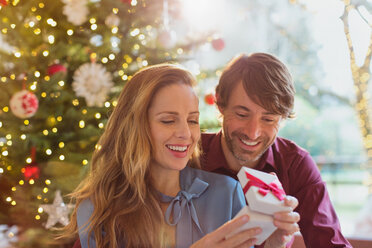 Ehemann beobachtet Frau beim Öffnen von Weihnachtsgeschenken vor dem Weihnachtsbaum - HOXF01974