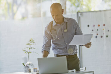 Male doctor with paperwork standing at laptop in doctor’s office - HOXF02016