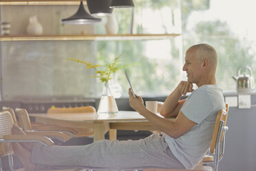 Älterer Mann, der sich entspannt und mit hochgelegten Füßen am Esstisch ein digitales Tablet benutzt - HOXF02048