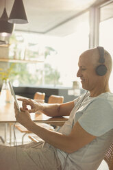 Älterer Mann, der mit Kopfhörern und digitalem Tablet am Esstisch Musik hört - HOXF02061