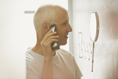 Älterer Mann rasiert sich mit elektrischem Rasierapparat vor einem Vergrößerungsspiegel im Badezimmer - HOXF02066