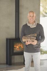 Porträt lächelnd reifen Mann hält Brennholz vor Holz brennenden Kamin - HOXF02073