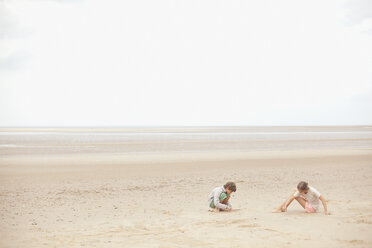 Brother and sister playing in sand on overcast summer beach - HOXF02183