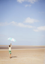 Junge hält gestreiften Regenschirm über Kopf am sonnigen Sommerstrand unter blauem Himmel - HOXF02184