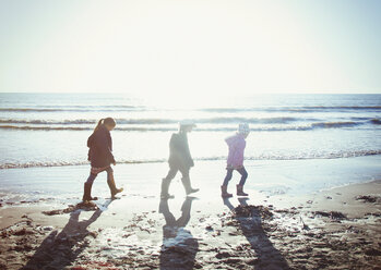 Brother and sisters in warm clothing walking in wet sand on sunny beach - HOXF02188