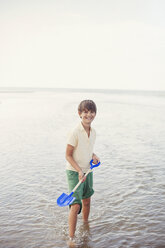 Portrait lächelnder Junge mit Schaufel in der Meeresbrandung am Sommerstrand - HOXF02189